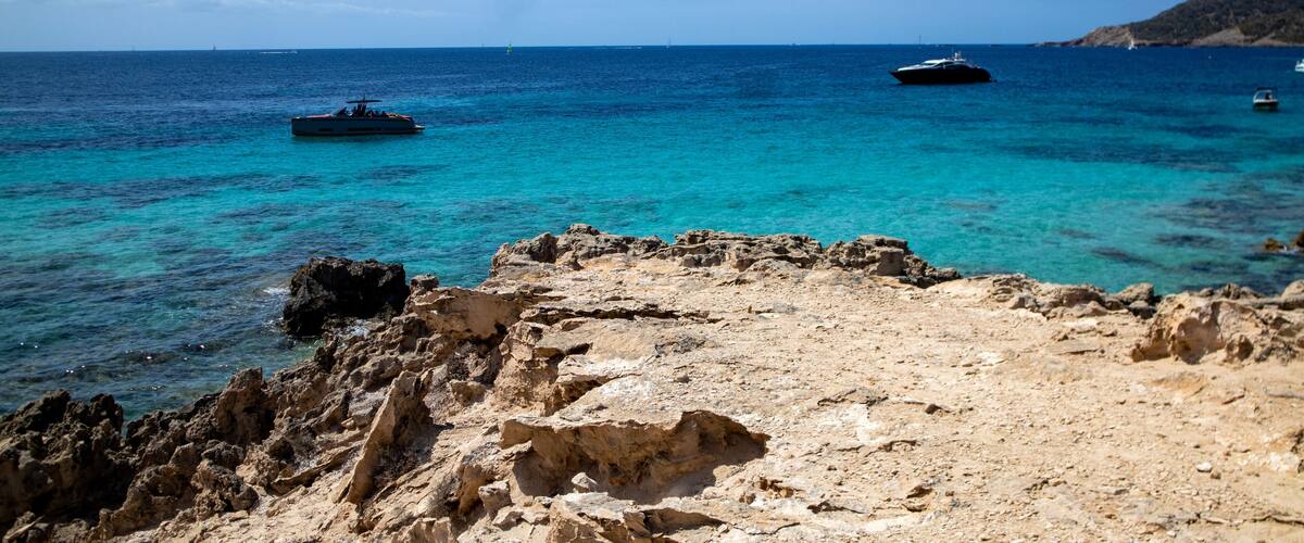 A rocky coastline with clear turquoise waters, boats, and a distant hill.