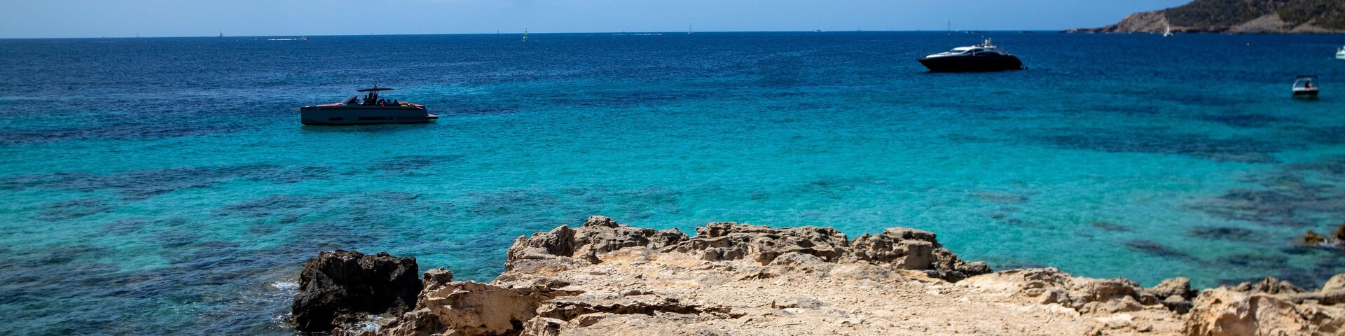 A rocky coastline with clear turquoise waters, boats, and a distant hill.