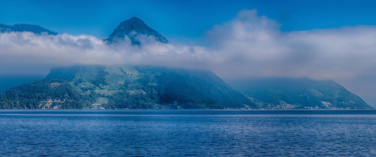 A mountain partially obscured by clouds, with a calm body of water in the foreground.