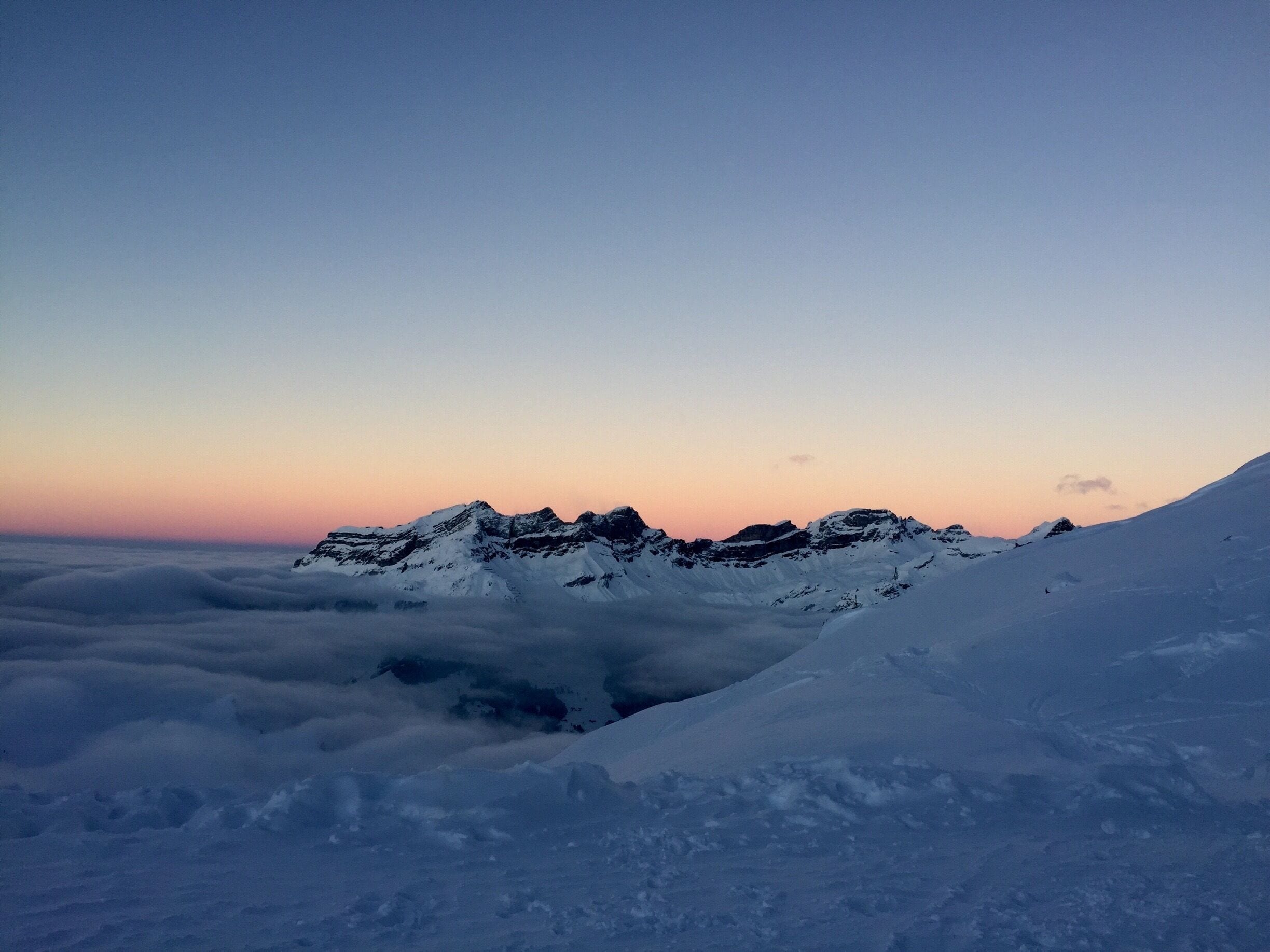 A snow-covered mountain range at sunset with clouds below.