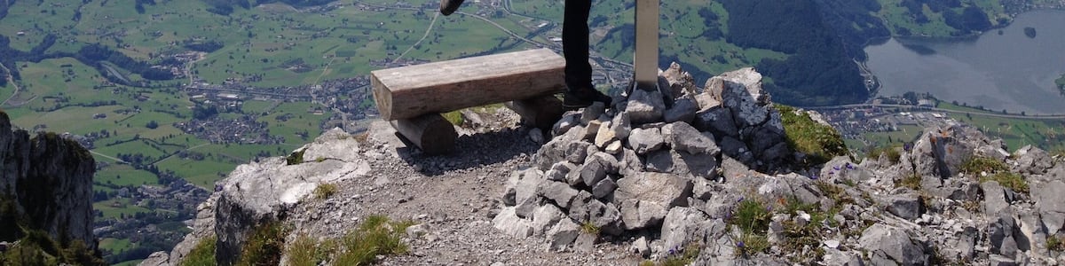 A person performing a yoga pose on a wooden bench at a mountain peak with a cross, overlooking a valley and a lake.