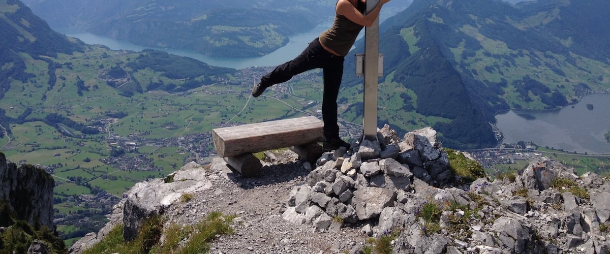 Una persona realiza una postura de yoga en un banco de madera en la cima de una montaña con una cruz, con vistas a un valle y un lago.
