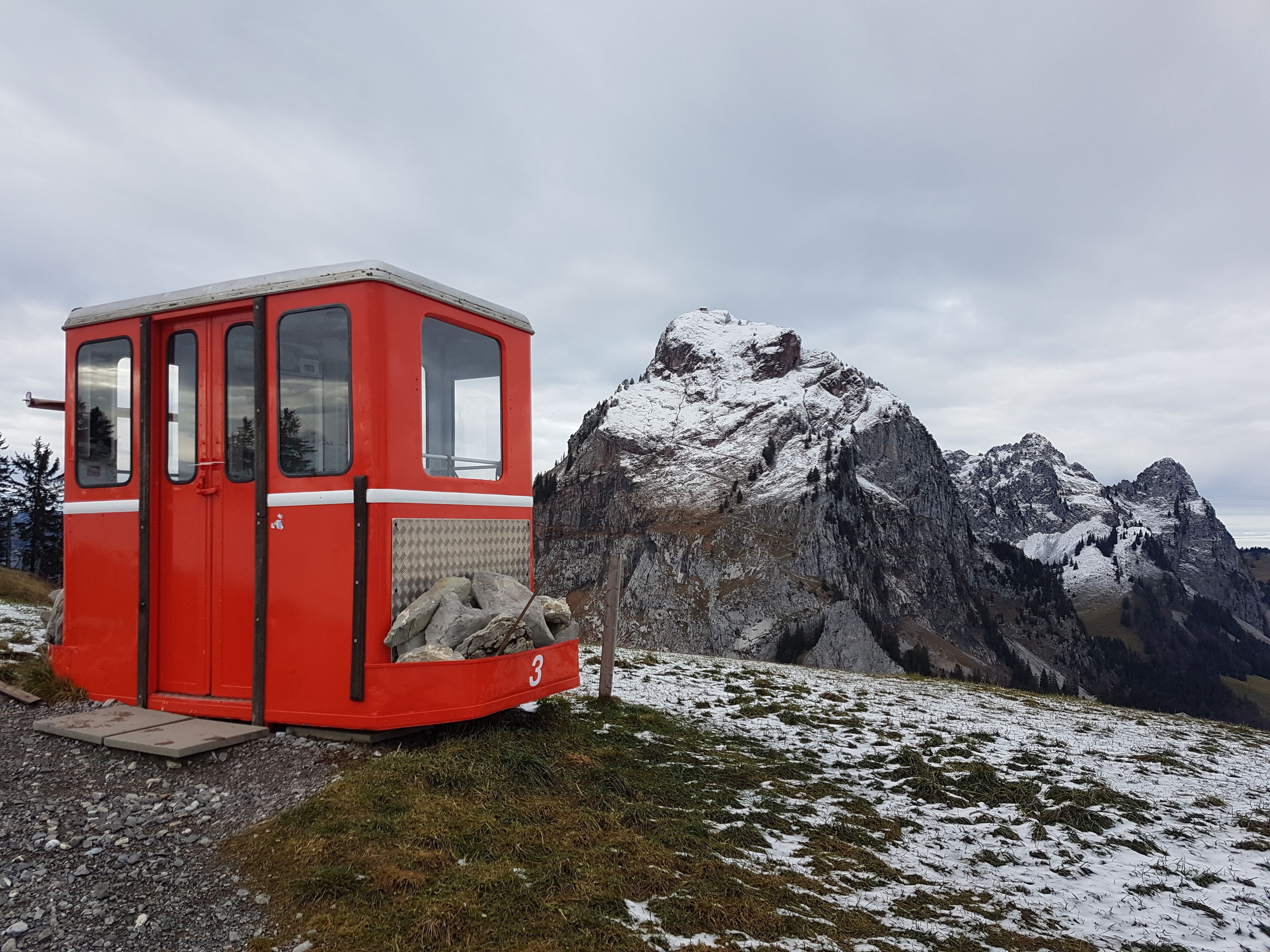 Una cabaña roja con el número 3 en su lateral, con montañas nevadas y un cielo nublado de fondo.