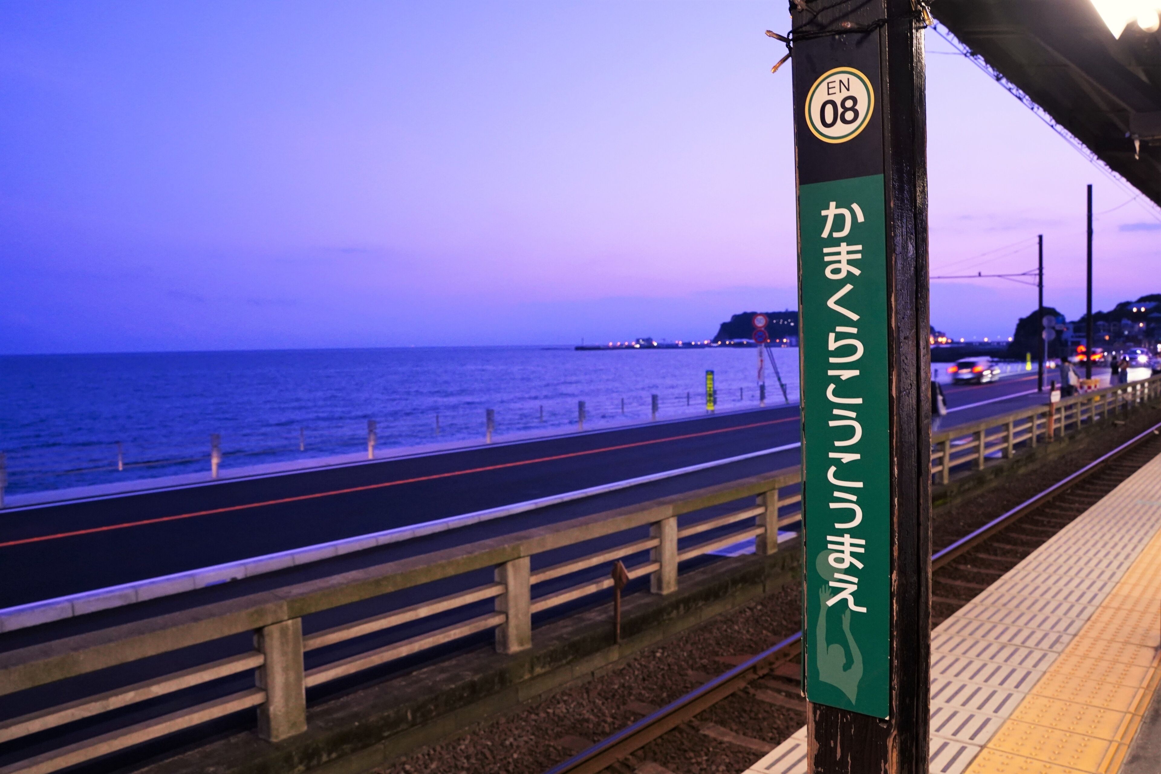 Andén de estación de tren costera, con un letrero en japonés, una vía de tren y vista al mar.