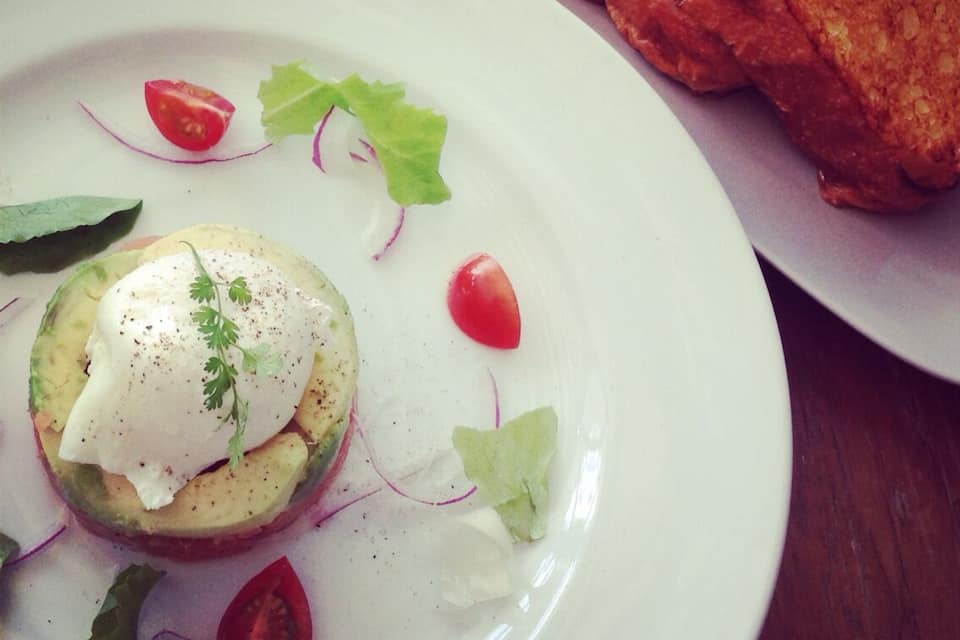 A plate with a poached egg on avocado, garnished with parsley, surrounded by cherry tomatoes and radish slices, and a side of toasted bread.