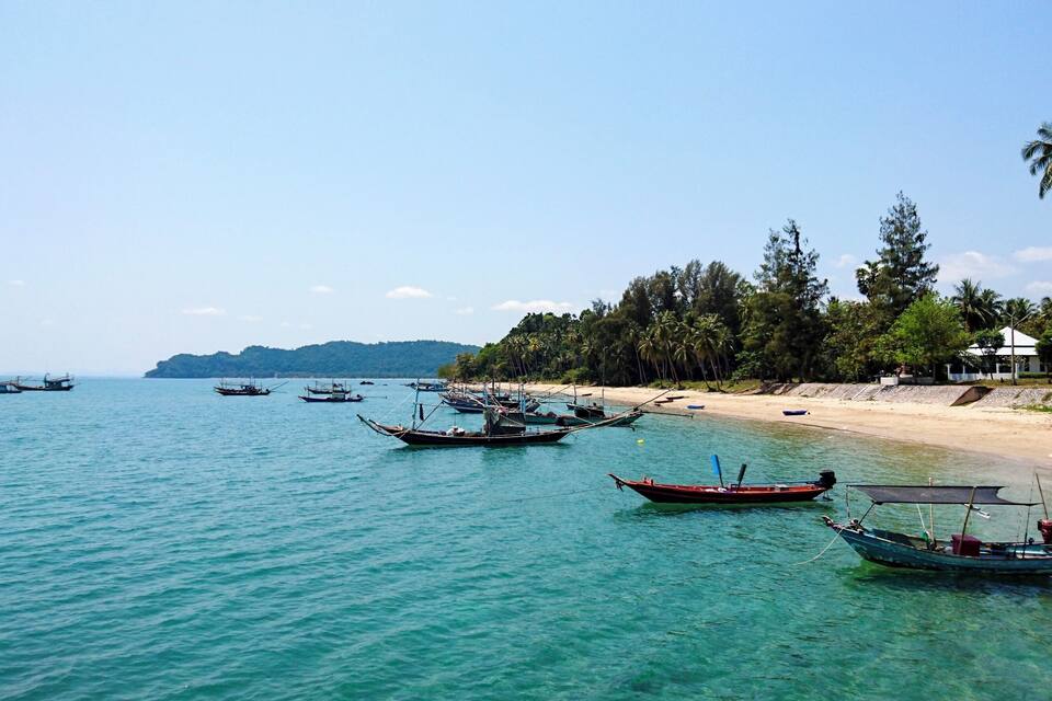 A beach with several boats anchored in the water, a sandy shore, and a line of palm trees.