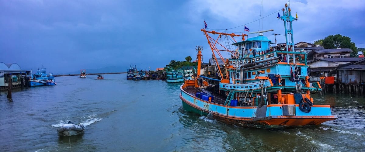 A harbor with several fishing boats docked, a small boat in the foreground, and a cloudy sky.