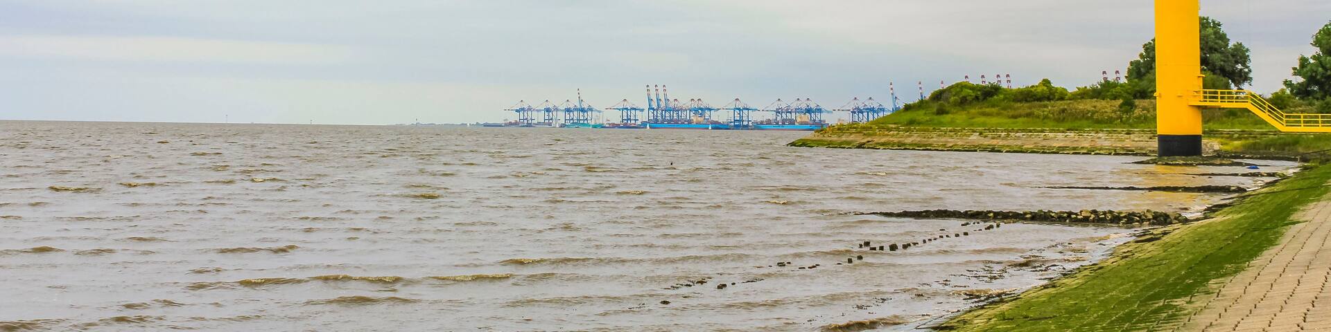 A yellow lighthouse by the sea, a blue crane structure in the distance, and a grassy embankment.