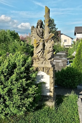 Une statue d’ange en pierre, ornée d’inscriptions, entourée d’une végétation luxuriante et de bâtiments résidentiels.