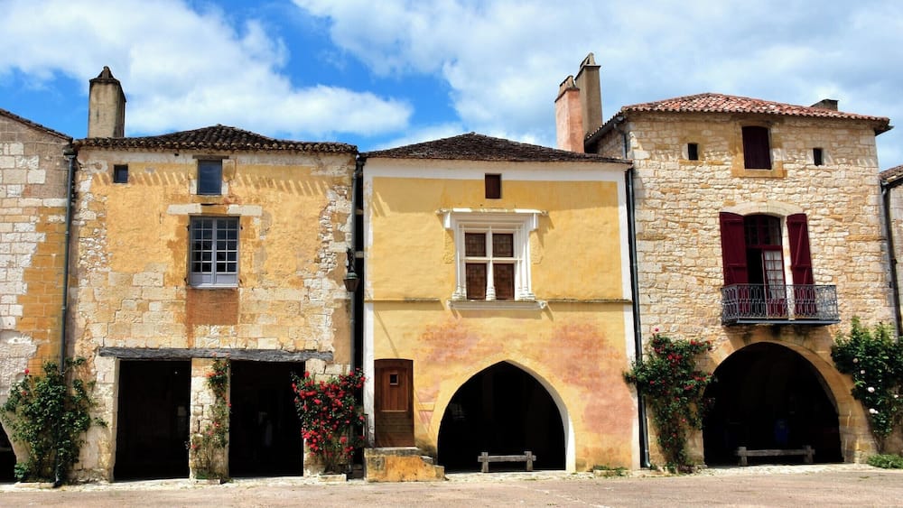 Une rangée de bâtiments historiques aux façades en pierre, aux portes en arc de cercle et aux volets rouges.