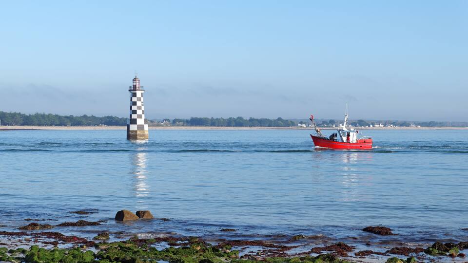 A lighthouse and a red boat on calm waters with rocky shorelines and a distant tree line.