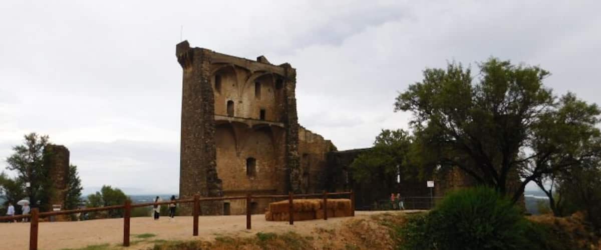 A historic stone tower with a rectangular base and a smaller upper section, surrounded by a fenced area and sparse vegetation.