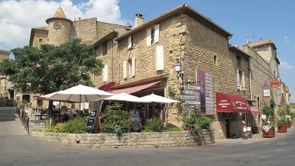 A stone building with a clock tower, outdoor seating with umbrellas, and various signs indicating directions and parking.