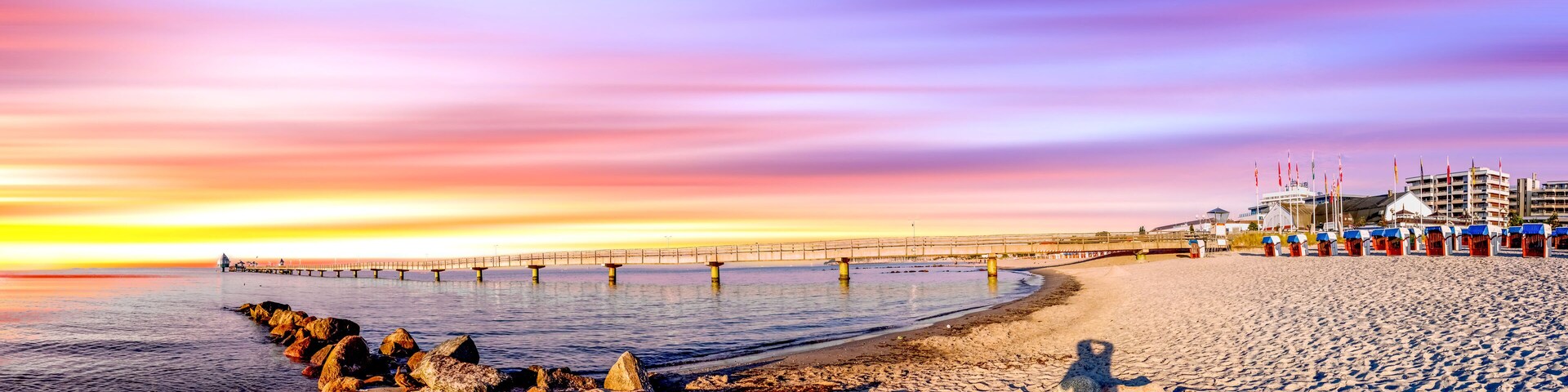 A beach with a pier extending into the water, a row of beach huts, and a sunset sky.