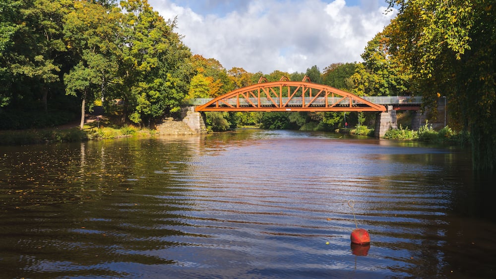 Eine rote Metallbrücke über einem ruhigen Fluss, umgeben von üppigen grünen Bäumen.