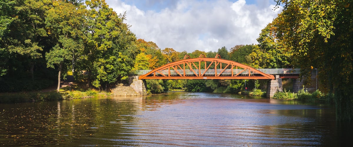 A red metal bridge over a calm river surrounded by lush green trees.