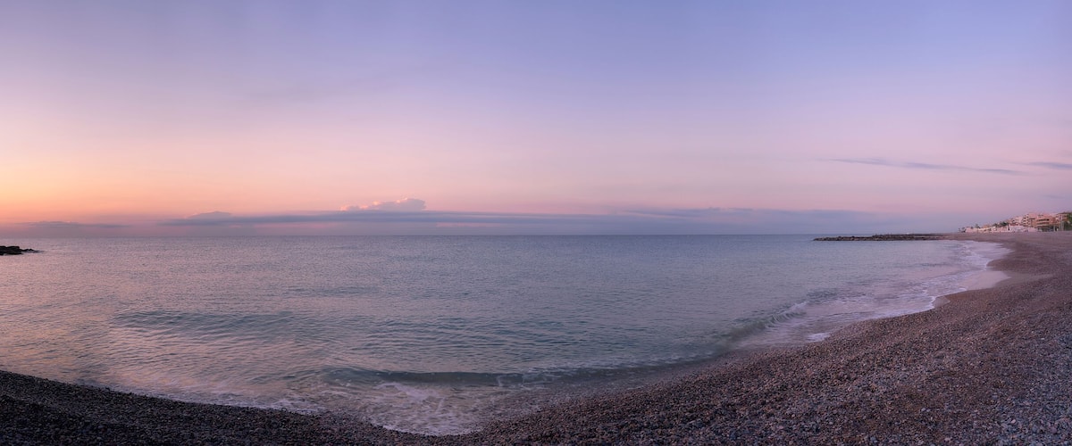 A pebbled beach at sunset with calm waters and a distant rocky outcrop.