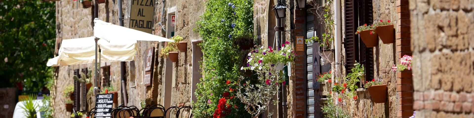 Una calle estrecha flanqueada por edificios antiguos, macetas con flores y un letrero de restaurante de pizza.