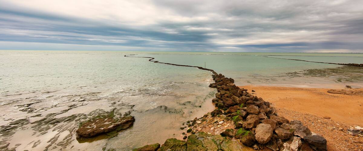 A coastal landscape with a sandy beach, rocky shoreline, and calm sea under a cloudy sky.