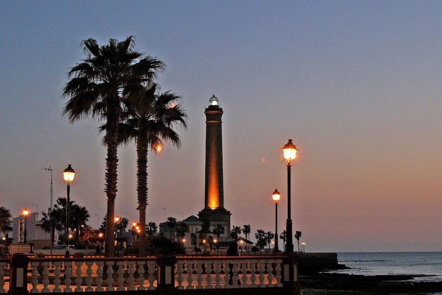 Un monumento costero con un obelisco alto, palmeras y farolas, sobre un cielo crepuscular.