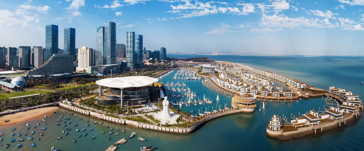 A coastal cityscape with modern skyscrapers, a marina with numerous boats, and a distinctive white statue.