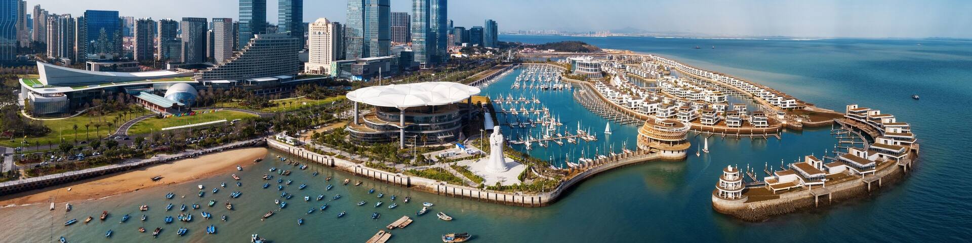 A coastal cityscape with modern skyscrapers, a marina with numerous boats, and a distinctive white statue.