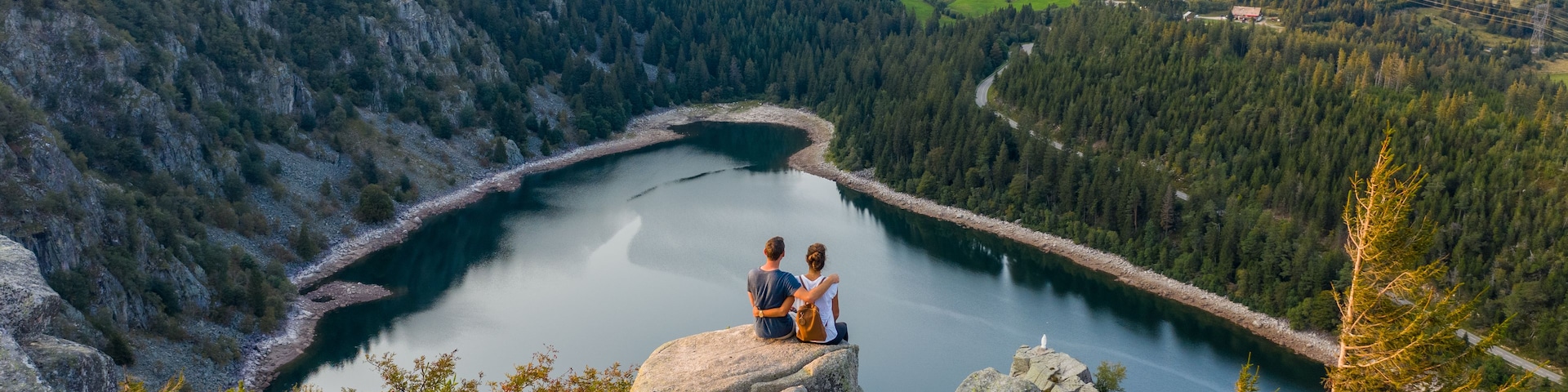 A lake surrounded by forested hills with a couple sitting on a rock overlooking it.