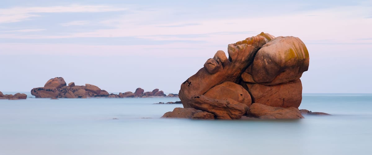 A large rock formation with a smooth, rounded shape, surrounded by calm water and distant smaller rocks.
