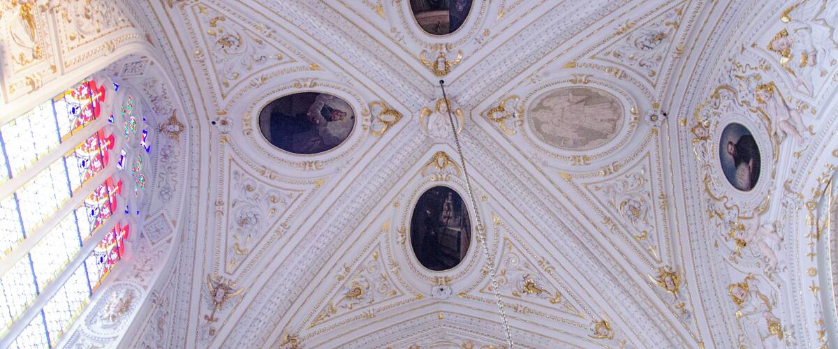 A ceiling with intricate gold and white patterns, multiple oval windows, and a stained glass window on the left.