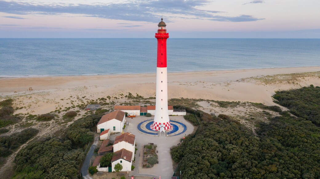 Paisagem costeira com um farol vermelho e branco imponente, uma praia de areia e vegetação ao redor.