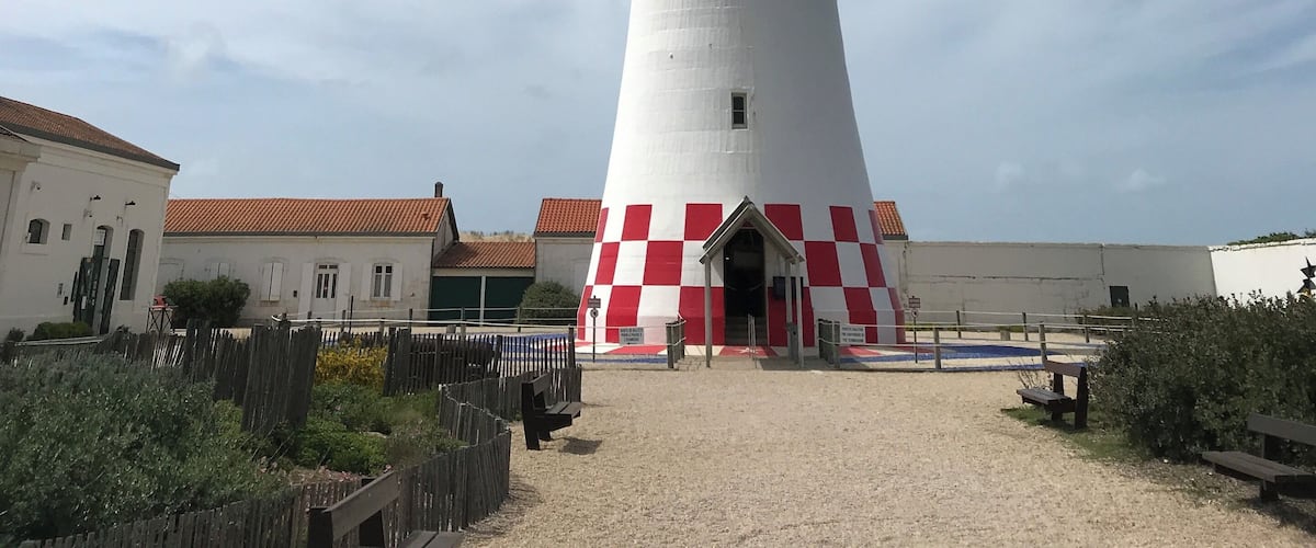 A tall red and white lighthouse with a checkered base, surrounded by a sandy area and buildings.