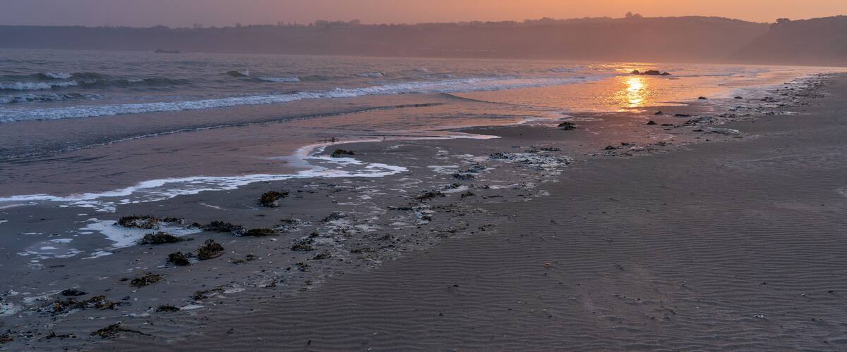 A beach at sunset with waves, a sandy shore, and distant hills.