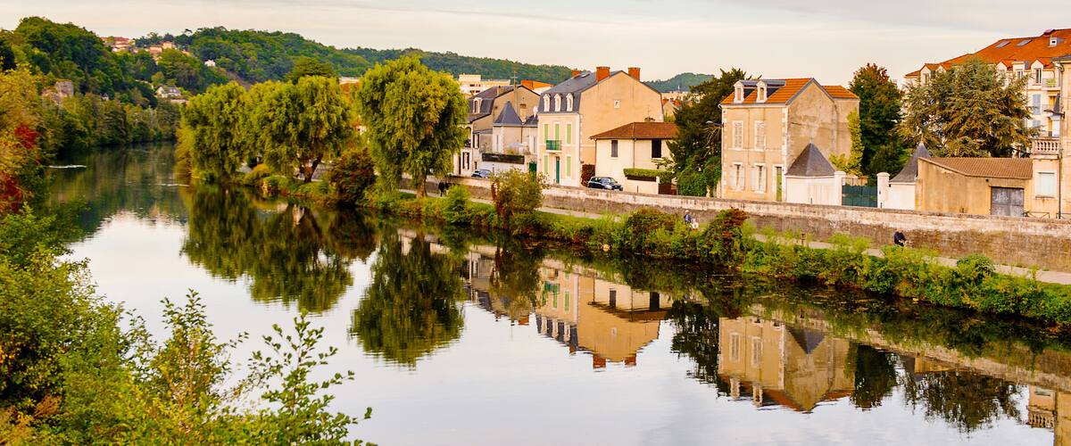 A calm river reflecting buildings and trees, surrounded by greenery and a clear sky.