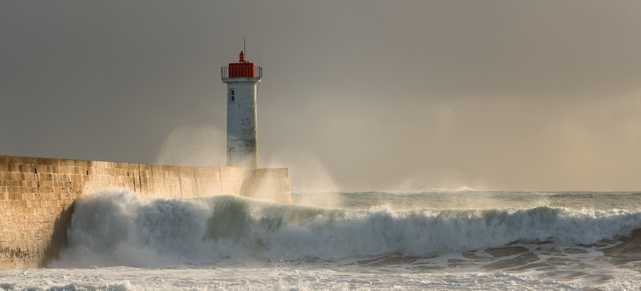 Un phare se dresse au bord d’une mer agitée, où les vagues s’écrasent contre un mur de béton.