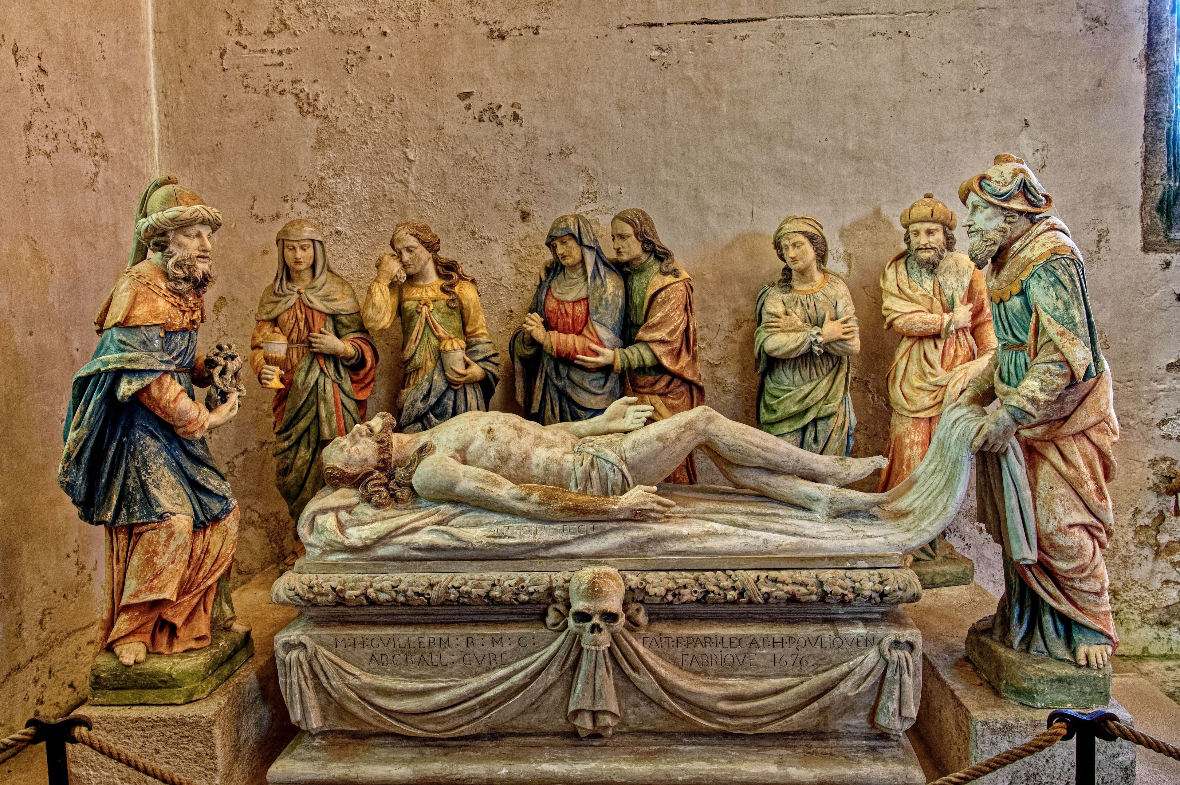 Une sculpture historique représentant une scène avec plusieurs personnages rassemblés autour d’une figure centrale allongée sur une civière, avec des inscriptions et un crâne à la base.