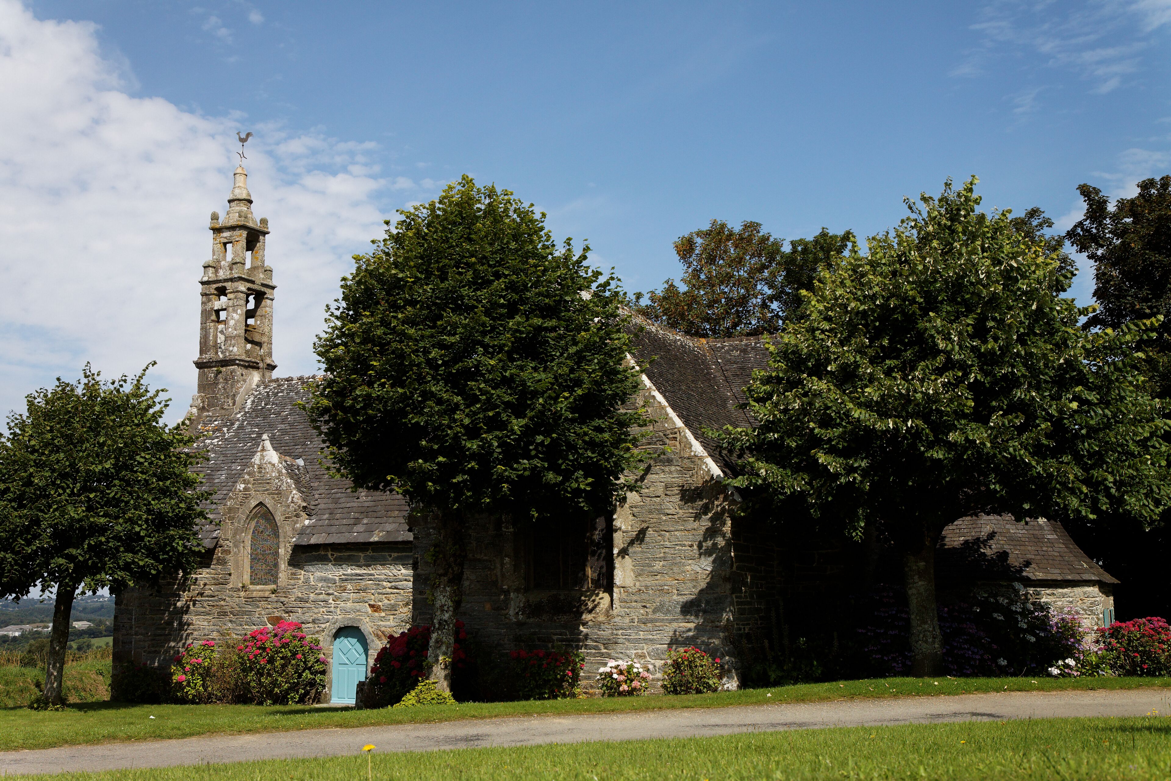 A stone church with a bell tower and a blue door, surrounded by lush greenery and flowering plants.