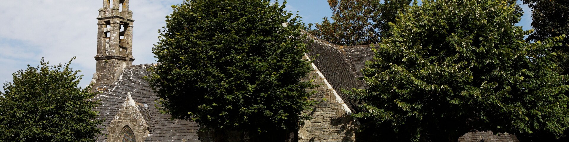 A stone church with a bell tower and a blue door, surrounded by lush greenery and flowering plants.