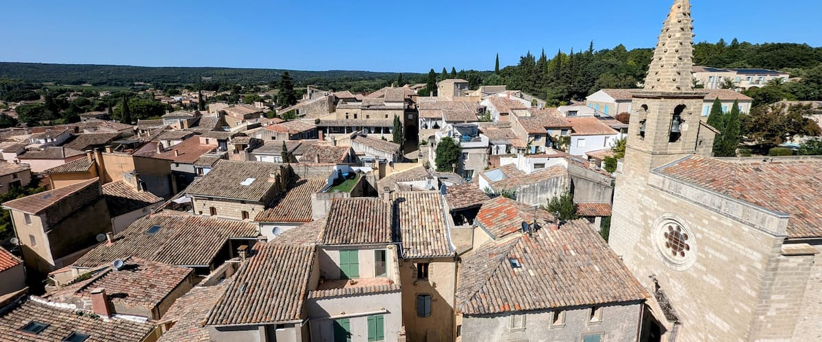 A historic town with terracotta-roofed buildings, a prominent church tower, and a lush green hillside in the background.
