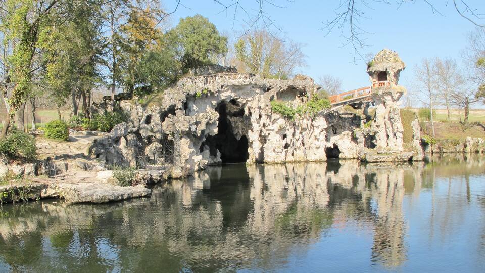 A rocky cave entrance by a calm pond, surrounded by trees and a stone bridge.