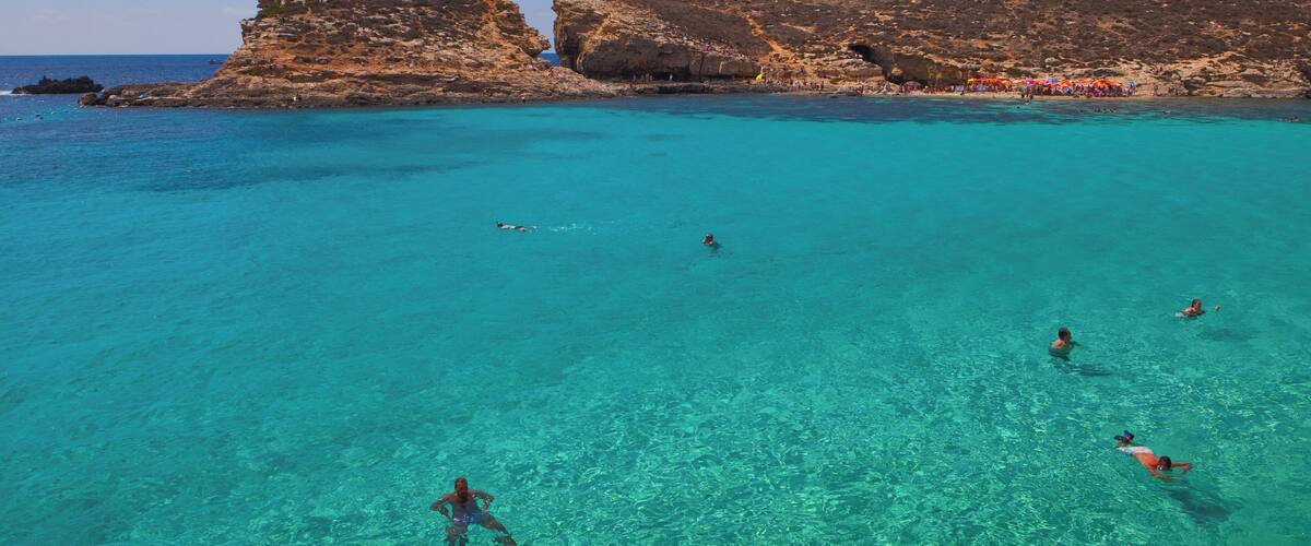 Une mer turquoise aux eaux limpides, bordée de falaises rocheuses, où l’on peut voir des gens se baigner.