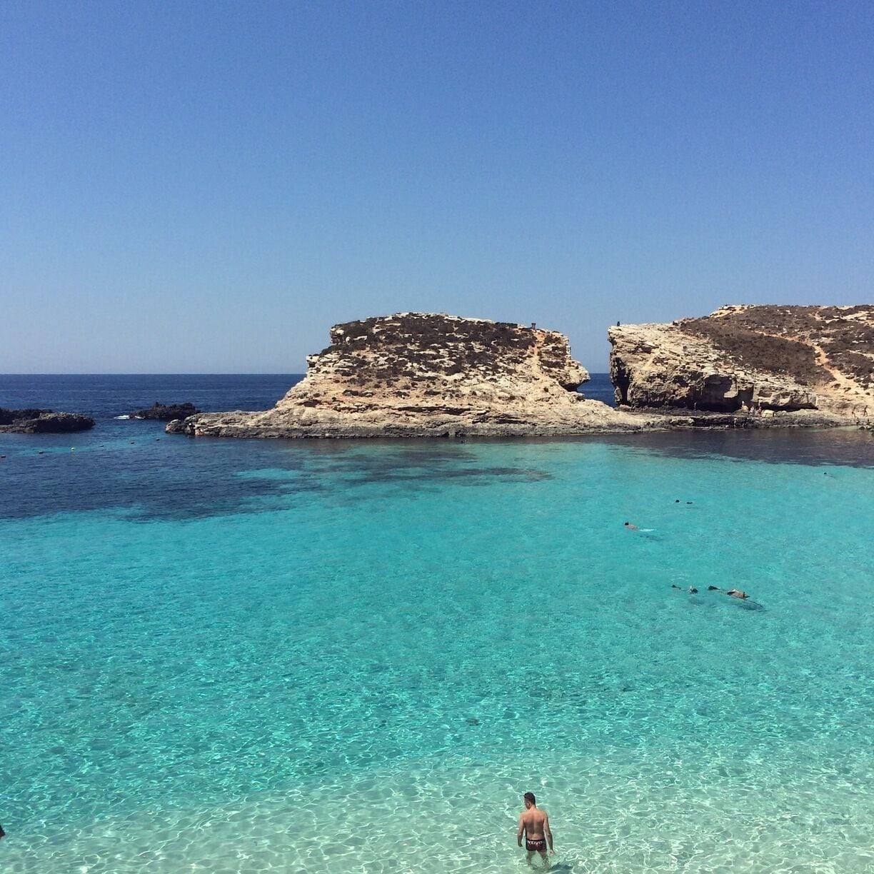 A clear turquoise sea with a person swimming, rocky cliffs, and a clear sky.