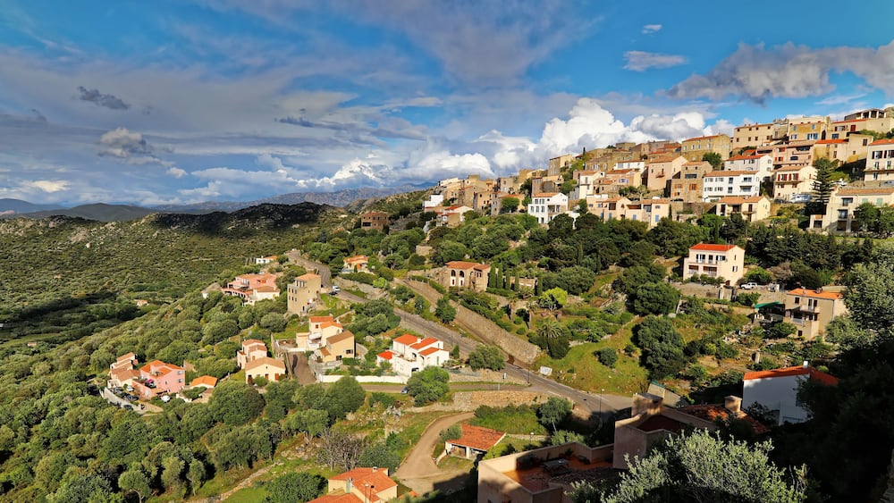 Un village perché sur une colline, avec des maisons à étages, une route sinueuse et une chaîne de montagnes en arrière-plan.