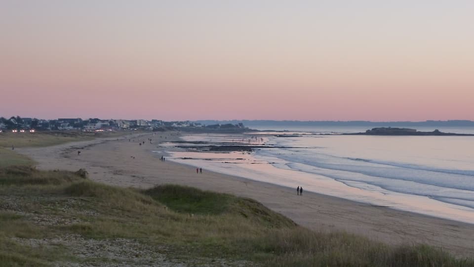 A beach at dusk with people walking along the shore and a distant cityscape.