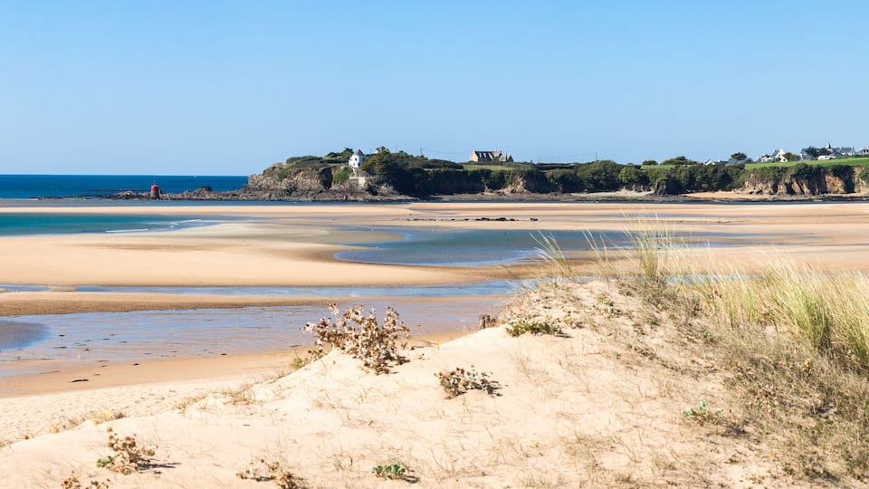 A coastal landscape with a sandy beach, tidal pools, and a lighthouse on a rocky outcrop.