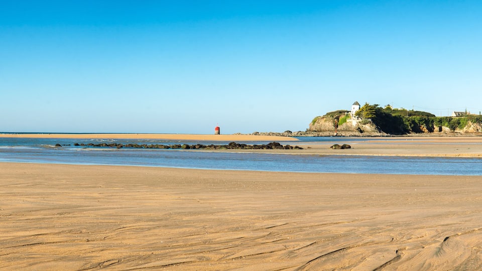 A sandy beach with a lighthouse, rocky outcrops, and a building on a cliff.