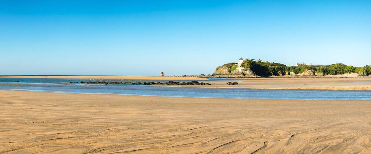 A sandy beach with a lighthouse, rocky outcrops, and a building on a cliff.