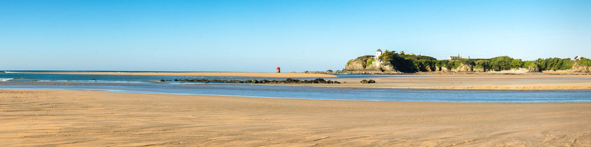 A sandy beach with a lighthouse, rocky outcrops, and a building on a cliff.