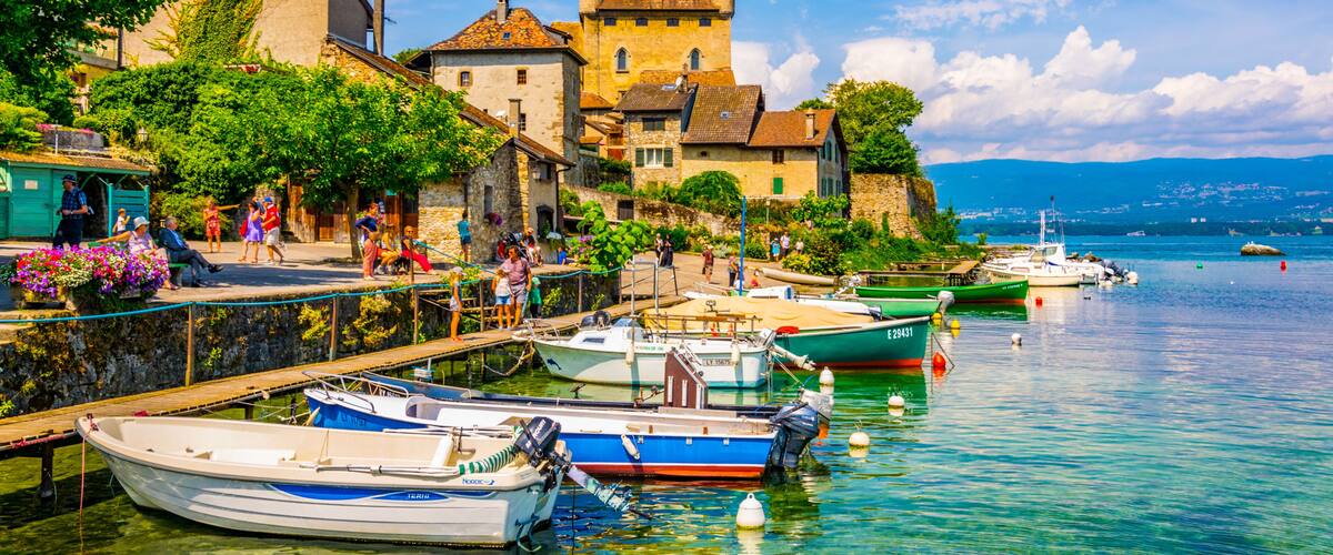 A picturesque lakeside village with historic buildings, a castle, and boats docked along the shore.