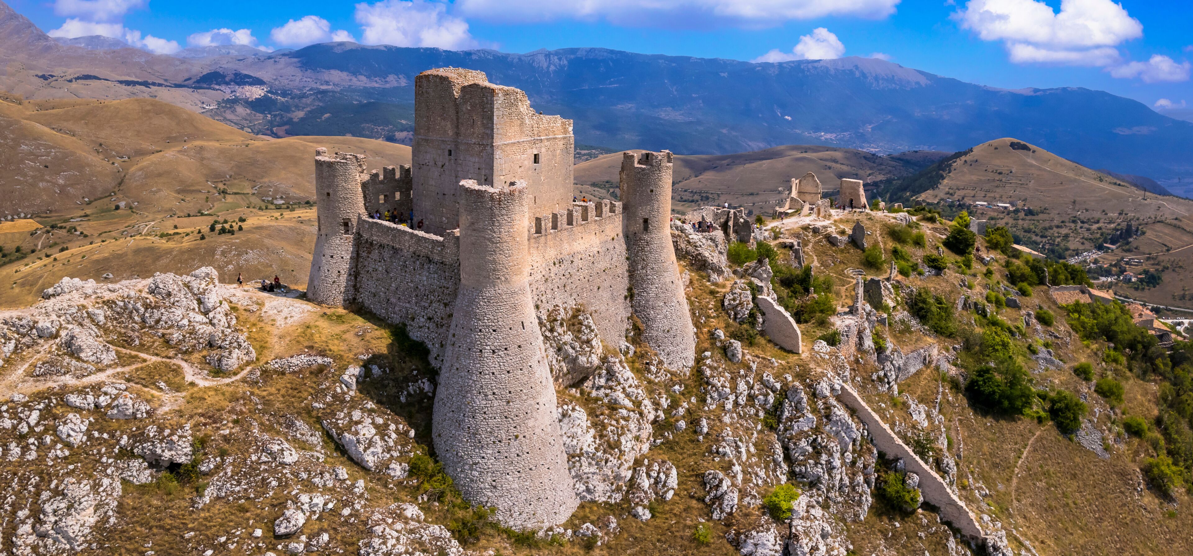 A medieval castle on a hill with surrounding mountains and sparse vegetation.