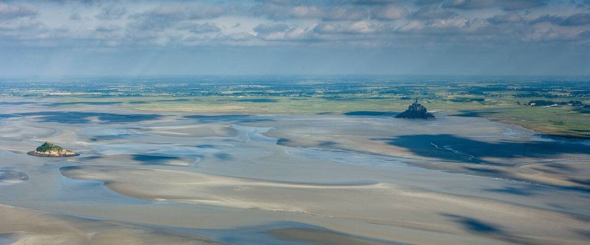 A coastal landscape with sandbanks, a small island, and a distant lighthouse.